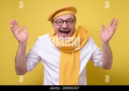 Surprised senior man in yellow scarf and beret holding hands up and smiling Stock Photo