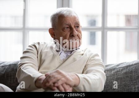 Pensive old man sitting on couch in living room alone Stock Photo