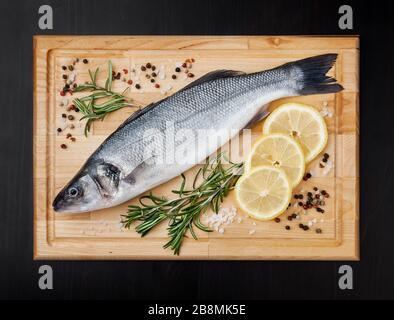 Fresh uncooked seabass with lemon, rosemary and spice on wooden board over black backdground Stock Photo