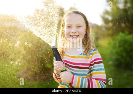 Adorable little girl playing with a garden hose on warm summer day. Child having fun with water on hot day. Outdoor summer activities for kids. Stock Photo