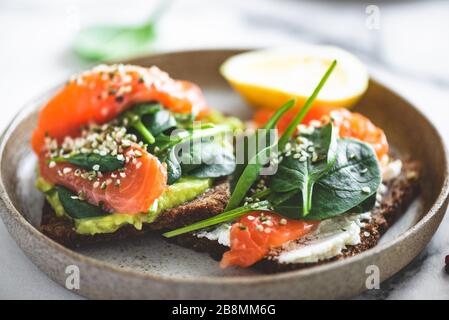 Rye bread toast with avocado, salmon, cream cheese, spinach leaf and hemp seeds on plate. Healthy food. Appetizer, breakfast or snack Stock Photo