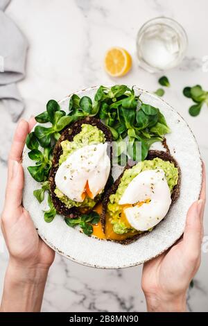 Avocado and poached egg on rye bread. Female hands holding healthy vegetarian breakfast or snack rye toast with mashed avocado, greends and egg Stock Photo