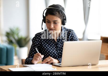 Focused Indian girl wearing headset writing notes, studying online Stock Photo