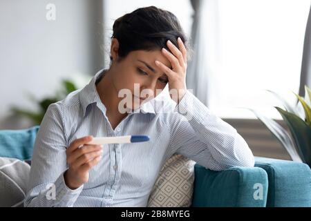 Unhappy Indian woman looking at pregnancy test, upset by result Stock Photo