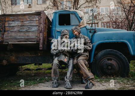 Couple in love sits in NBC protective suits and gas mask near old truck. Concept of a preventive measures and protection for coronavirus COVID 19 pand Stock Photo