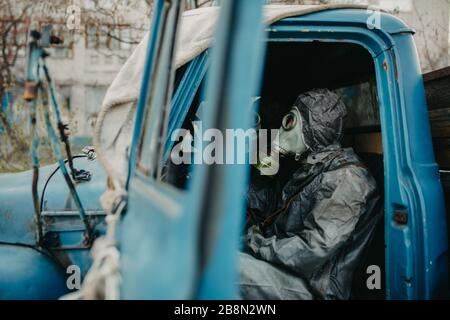 Two persons sits in NBC protective suits and gas masks in old truck. Concept of a preventive measures and protection for coronavirus COVID 19 pandemic Stock Photo