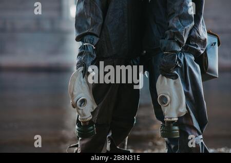 Couple in love stands under bridge in NBC protective suits and gas masks in their hands. Concept of a preventive measures and protection for coronavir Stock Photo