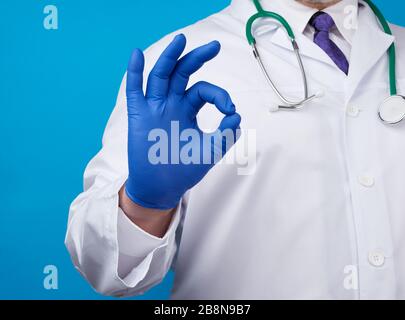 male doctor in white coat, blue latex medical gloves shows ok gesture with his right hand, concept of hope that everything will be fine, blue backgrou Stock Photo