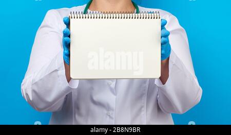 medic woman in white coat, blue latex gloves holding an open notebook with blank white sheets, blue background, place for text Stock Photo