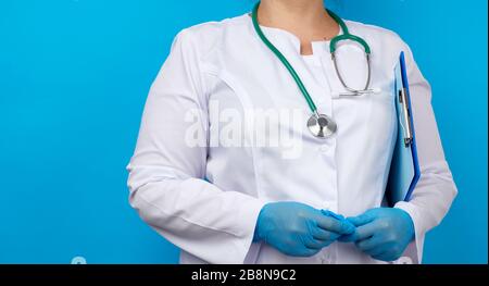 medic woman in white coat, blue latex gloves holding a tablet with a clip for papers, blue background, place for an inscription Stock Photo