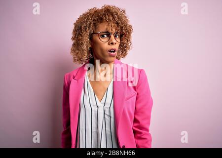 Young african american businesswoman wearing glasses standing over pink background In shock face, looking skeptical and sarcastic, surprised with open Stock Photo