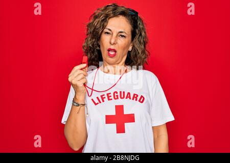 Middle age senior summer lifeguard woman holding whistle over red background In shock face, looking skeptical and sarcastic, surprised with open mouth Stock Photo