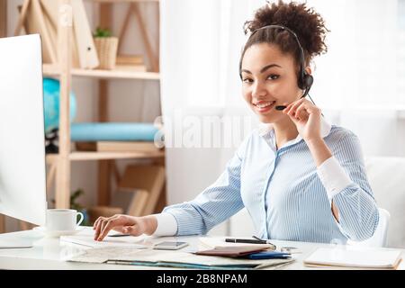 Tour agent in headset sitting at table in office holding mike looking camera happy using computer Stock Photo