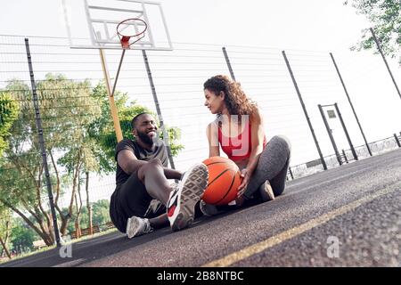 Outdoors Activity. African couple sitting with ball on basketball court talking laughing cheerful Stock Photo