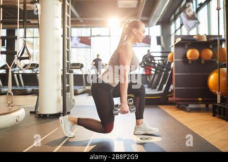 Full length side view portrait of sportive young woman doing squats with weights during workout in sunlit gym, copy space Stock Photo
