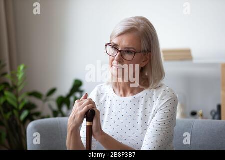 Pensive older lady in eyeglasses sitting on couch with cane. Stock Photo