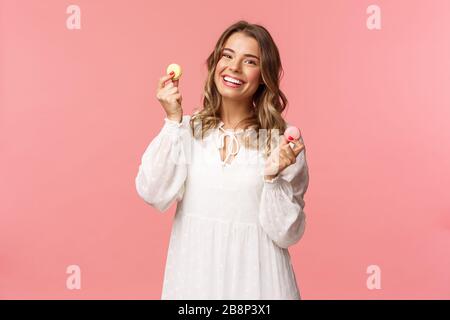 Holidays, spring and party concept. Portrait of tender, lovely blond woman in white dress, dancing joyfully with two macarons, smiling happy eating Stock Photo