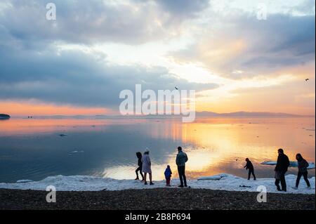 Silhouettes of people walking by the sea with the edge of the ice and admiring the incredibly beautiful orange and blue sunset with clouds and rays of Stock Photo