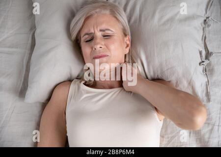 Older unhappy woman lying in bed, feeling strong neck ache. Stock Photo