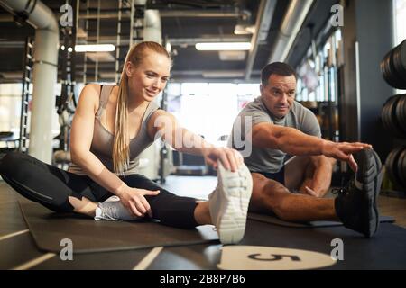 Full length portrait of smiling sportive couple doing stretching exercises while sitting on floor in gym and enjoying workout Stock Photo