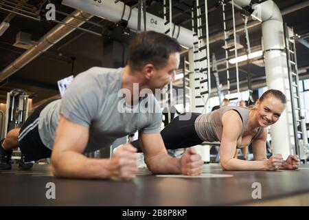 Full length portrait of cheerful sportive couple doing plank exercises during workout in modern gym and looking at each other, copy space Stock Photo