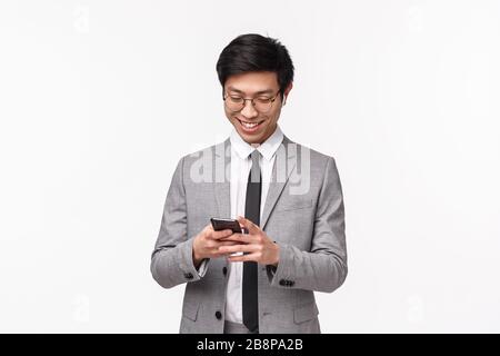 Waist-up portrait of handsome successful young asian male entrepreneur using mobile phone, texting girlfriend, smiling at smartphone display, watching Stock Photo