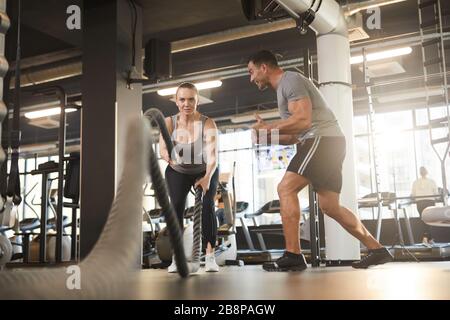 Full length portrait of young woman doing battle rope exercises during cross workout in modern gym with personal coach motivating her, copy space Stock Photo