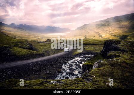 white camper van driving along winding mountain road through beautiful sunlit valley surrounded by mountains, Iceland Stock Photo