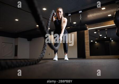 Full length wide angle portrait of focused young woman doing battle rope exercises during cross workout in dark gym, copy space Stock Photo