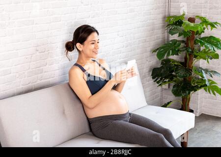 Happy Chinese pregnant female in sportswear smiling and examining sonogram while sitting on sofa in cozy living room Stock Photo