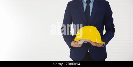 A businessman stands and holds a yellow helmet in his hand and behind him is a white wall. Stock Photo