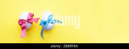 Two white eggs wrapped in blue and pink ribbons, on a yellow background. Banner. Easter concept. Stock Photo