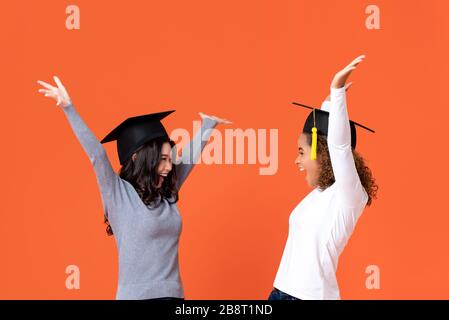 Happy excited young female students wearing graduate caps smiling with hands raising celebrating graduation day isolated on orange background Stock Photo
