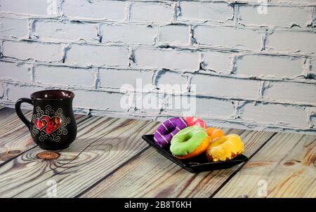A rectangular plate with five multi-colored donuts and a black ceramic coffee mug stand on a wooden table. Close-up. Stock Photo