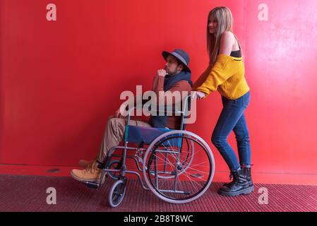 Disabled in a wheelchair pushed by a young blonde girl in front of a red background wall Stock Photo