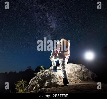 Back view of space traveler climbing up rocky cliff under beautiful night sky with stars. Cosmonaut in space suit exploring new planet. Concept of space travel, galaxy and human in cosmos.