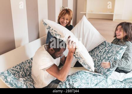 happy multi-ethnic family has fun in the on the bed doing a pillow fight, African dad attacked by his Caucasian wife and daughter, they enjoy the time Stock Photo