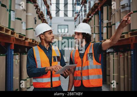 Tensed worker in factory showing shelves to his manager holding digital tablet with hardhat and safety vest Stock Photo