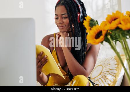 Smiling female student listening to music in her headphones when reading book and getting ready for class Stock Photo