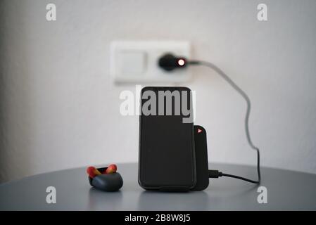 Black desktop wireless charger with charging led charging a smartphone. Display in black. Silver gray table. Ear plugs on bedside table Stock Photo