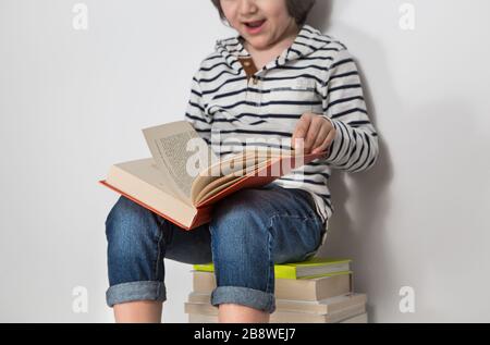 Preschooler sitting on a pile of books while leafing through a red book Stock Photo