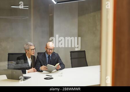 Wide angle portrait of two successful mature business people reading notes while sitting at table in minimal office interior behind glass wall, copy s Stock Photo