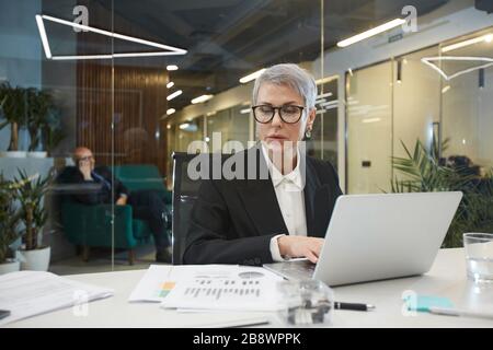 Portrait of successful mature businesswoman using laptop while working with documents at desk in glass office, copy space Stock Photo