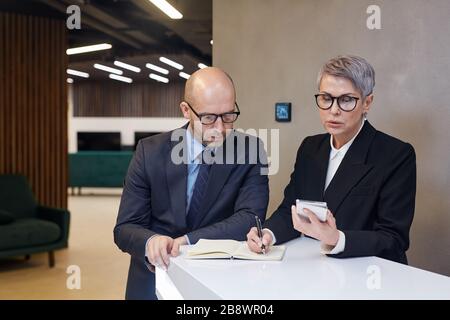 Portrait of two mature business people writing out data from smartphone at stand in minimal office interior, copy space Stock Photo