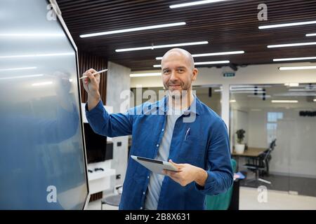 Waist up portrait of bald adult man smiling at camera while using digital board during presentation or training seminar in office, copy space Stock Photo