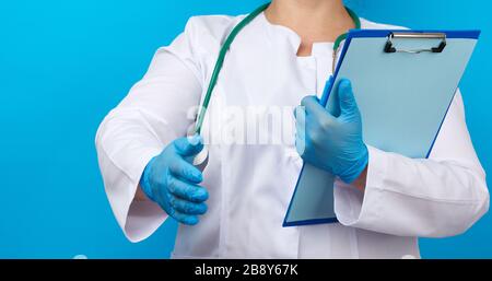 medic woman in white coat, blue latex gloves holding a tablet with a clip for papers, right hand pulls forward for a handshake, blue background Stock Photo