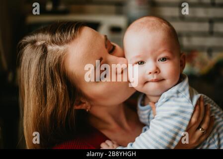 A mom holds a child in her arms. A mother kisses a toddler. A young women hugs her infant. A girl has fun and rejoices with the baby.Close up. Stock Photo