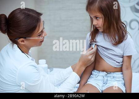 Nice woman doctor measures the temperature of a baby girl, little kid with a thermometer, scheduled examination at the pediatrician's office Stock Photo