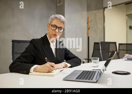 Minimal portrait of successful mature businesswoman working while sitting against grey wall at workplace, copy space Stock Photo