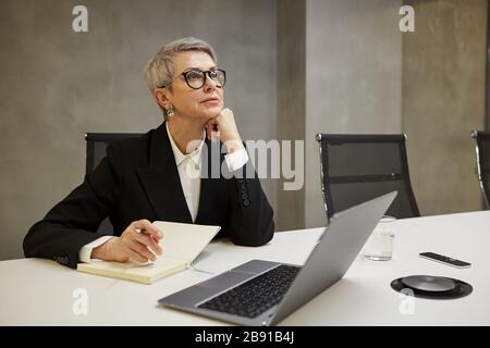 Minimal portrait of successful mature businesswoman looking away pensively while sitting against grey wall at workplace, copy space Stock Photo